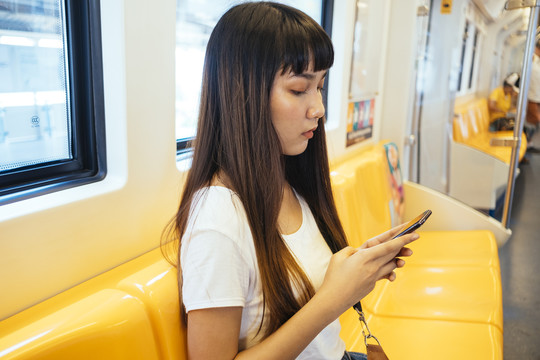 一个留着刘海发型的亚洲女孩坐在火车上的黄色长凳上玩手机的特写镜头。