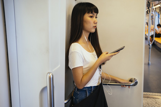 可爱的亚洲女孩在上班时玩智能手机，戴着耳机听音乐。在火车上发生的。