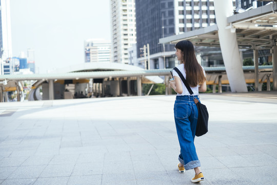 穿着白色衬衫和蓝色牛仔裤的年轻亚洲女人在城市里散步。城市旅游概念。