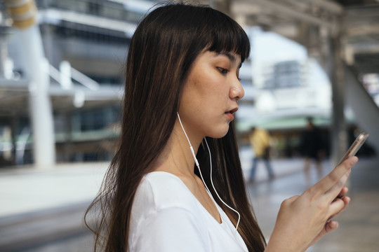 刘海发型的年轻亚洲女性侧视用耳机听手机里的音乐。