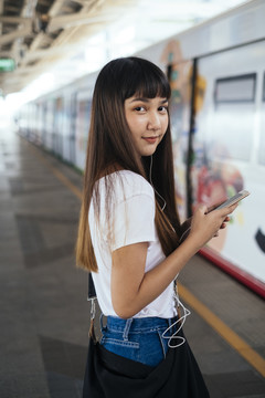 站在火车站台上，用智能手机做刘海发型的可爱泰国年轻女子。