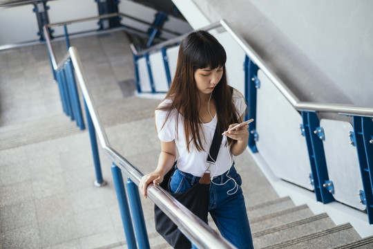 穿着白色衬衫和蓝色牛仔裤的可爱泰国年轻女子使用智能手机走上楼梯。