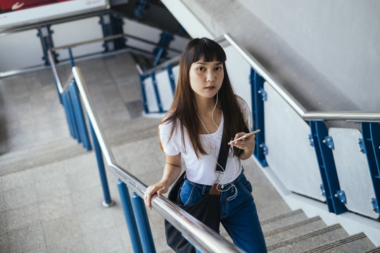 穿着白色衬衫和蓝色牛仔裤的可爱泰国年轻女子使用智能手机走上楼梯。