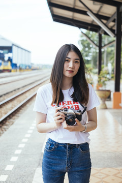 在火车站月台上，一位年轻的亚裔泰国长毛黑发女旅行者手持相机。