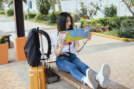 年轻的亚裔泰国长毛黑发女旅行者坐在火车站的长凳上看地图。