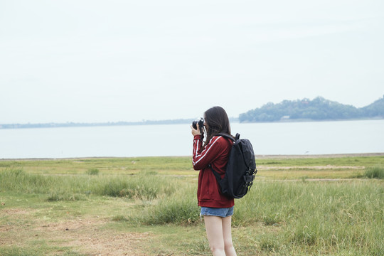 在春武里的湖边，一位年轻的亚裔泰国长发旅行者，身穿红色夹克，手持相机。