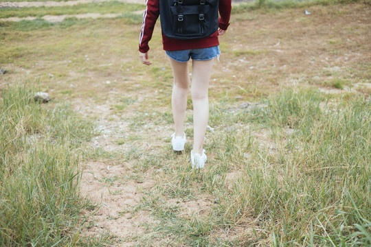 后视图-年轻的亚泰黑头发旅行家妇女走在绿色田野的乡村边。复制空间。