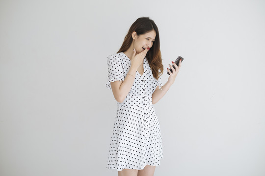 一位穿着黑白圆点连衣裙的年轻泰国女士手持智能手机，看着手机上的购物促销。在白色背景上隔离。