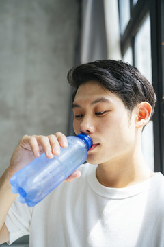 穿着白衬衫的亚洲年轻人在家里喝瓶装水。