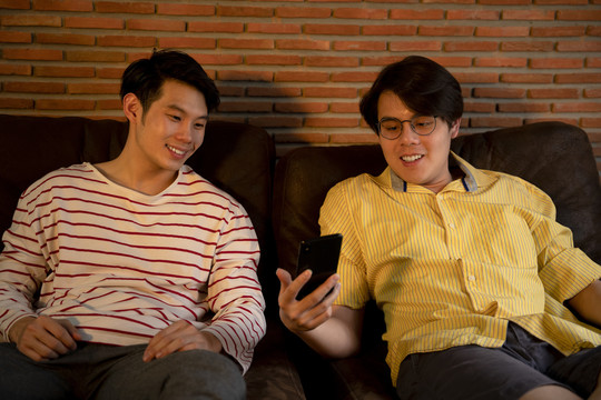 一对亚洲LGBTQ+男士坐在沙发上，一边喝酒一边看着智能手机庆祝。