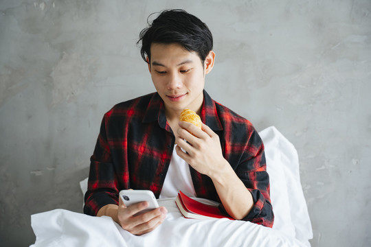 亚洲年轻人在床上吃早餐和使用智能手机。