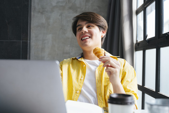 穿着黄衬衫的亚洲年轻人在现代办公室工作和吃零食。