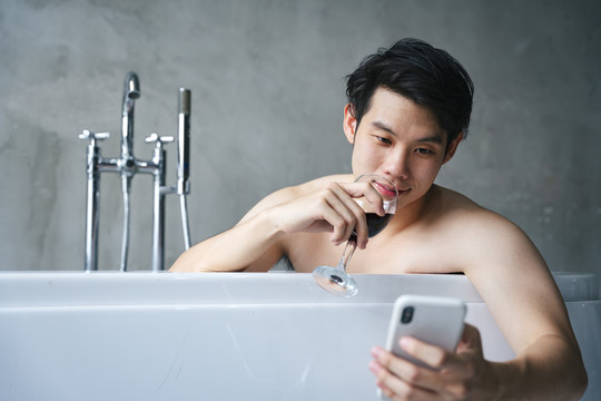 亚洲年轻人在浴缸里喝酒，使用智能手机。