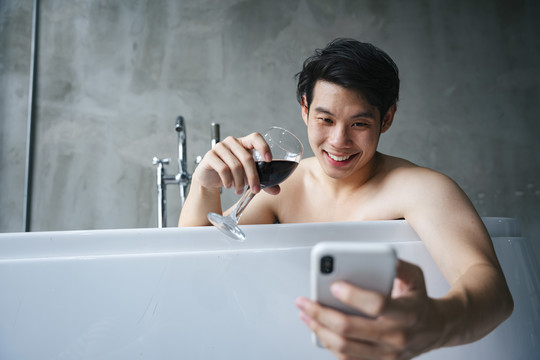 亚洲年轻人在浴缸里喝酒，使用智能手机。