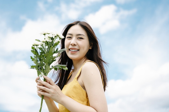 穿着黄色衣服的美丽亚洲女人的肖像，在夏天的云彩背景上举着白色的花。