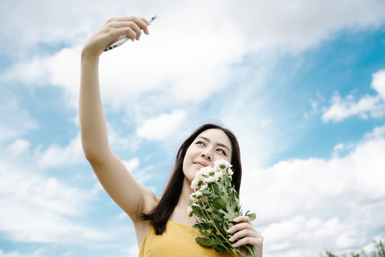 美丽的亚洲女子手持白色花朵在田野上用智能手机自拍。