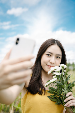 美丽的亚洲女子手持白色花朵在田野上用智能手机自拍。