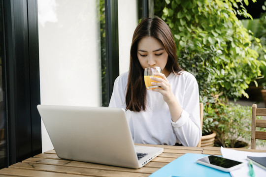 一位穿着白衬衫的美丽亚洲女性，一边用笔记本电脑工作，一边喝着橙汁。