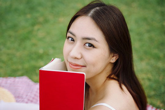 美丽的亚洲旅行家女子拿着红皮书躺在户外公园的垫子上。