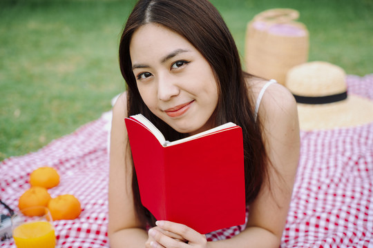 美丽的亚洲旅行家女子拿着红皮书躺在户外公园的垫子上。