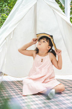 可爱的亚洲小女孩，头上戴着一本书，呆在花园里的白色露营帐篷里。