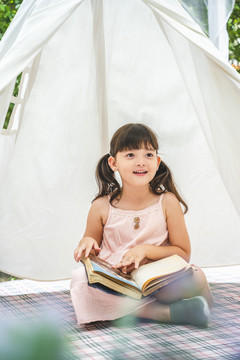 可爱的亚洲小女孩在户外花园的白色露营帐篷里看书。