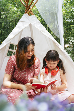 母亲和女儿呆在白色的露营帐篷里，在户外花园里读着一本书中的故事。