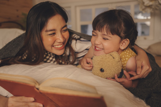 亚洲的母亲和女儿晚上在卧室里读床单故事书。