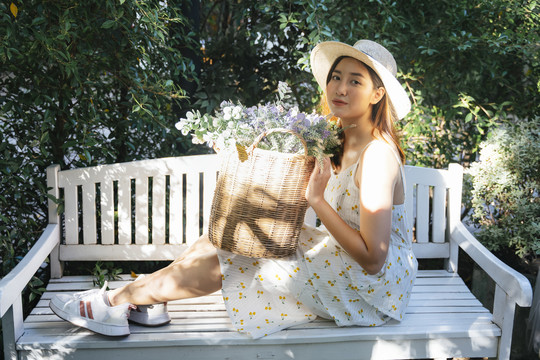 美丽的亚洲女人戴着帽子，穿着白色的衣服，花篮坐在花园里的白色长凳上。