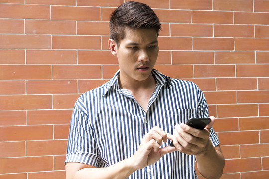 一名亚洲男子站在砖墙后面，用智能手机做网上银行业务。