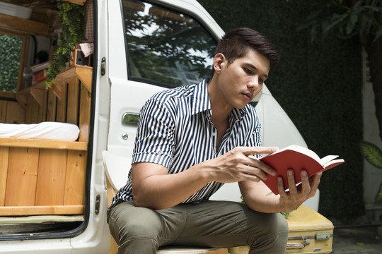 一名亚裔男子在户外露营地的CV车旁看书。