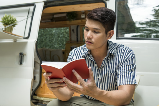 一名亚裔男子在户外露营地的CV车旁看书。