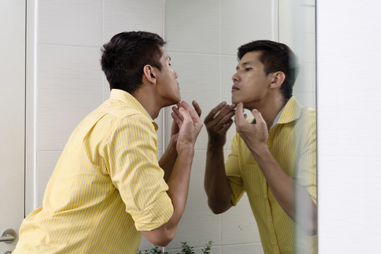 亚洲男人对着镜子检查面部痤疮问题。
