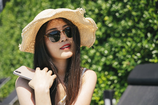 一位年轻的亚洲旅行家女士的特写照片，她在户外戴着墨镜，戴着帽子，手持智能手机。