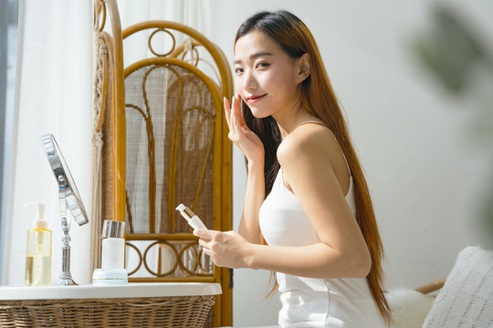 侧视图-美丽的年轻亚洲女性在自然光下在家中涂抹乳液。