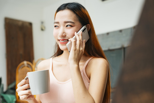 美丽的亚洲年轻女子，在卧室里端着一杯咖啡和智能手机。