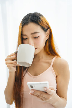 美丽的亚洲年轻女子，手拿咖啡，在白色背景下使用智能手机。