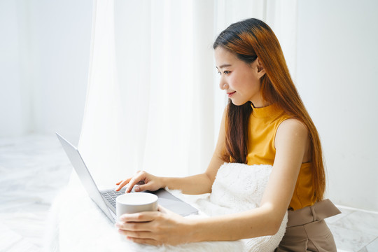 一位年轻的亚洲女性正在使用笔记本电脑，手里拿着一杯咖啡。