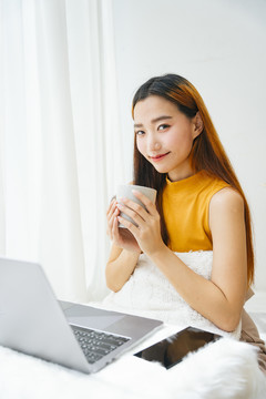 美丽的亚洲年轻女子在笔记本电脑桌前端着一杯咖啡。
