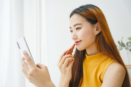 年轻漂亮的亚洲女子涂唇膏，在智能手机上看镜子。