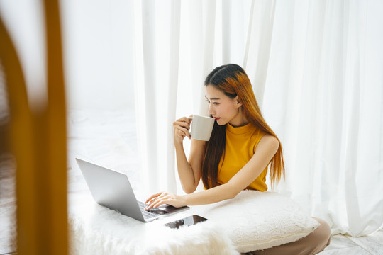 一位年轻漂亮的亚洲女士，一边喝着咖啡，一边用笔记本电脑工作。