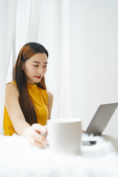 一位年轻漂亮的亚洲女士，手里拿着一杯咖啡，看着笔记本电脑。