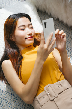 美丽的亚洲年轻女子在家里用智能手机躺在枕头上。