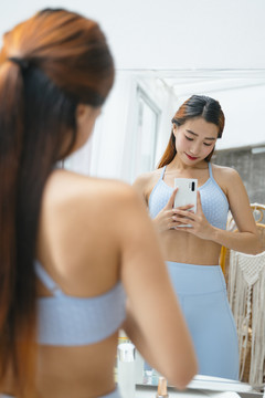 穿着运动胸罩的美丽运动型亚洲女性在镜子前拍照。她真为自己的身体感到骄傲。