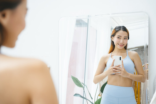 穿着运动胸罩的美丽运动型亚洲女性在镜子前拍照。她真为自己的身体感到骄傲。
