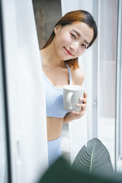 美丽的运动型亚洲女性，穿着运动胸罩，在家阳台上端着一杯咖啡。