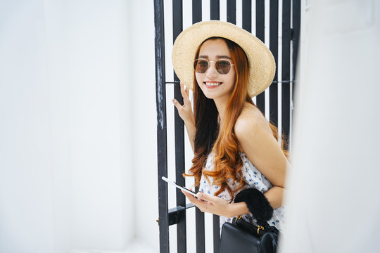 美丽的亚洲女旅行者戴着太阳镜，戴着帽子，手持智能手机在酒店门口。