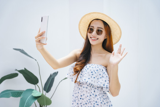 美丽的亚洲女旅行者戴着太阳镜，戴着帽子，用智能手机观看视频或直播。