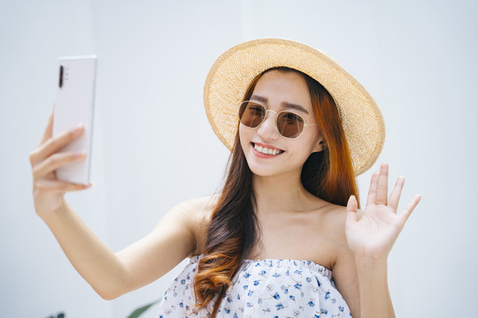 美丽的亚洲女旅行者戴着太阳镜，戴着帽子，用智能手机观看视频或直播。