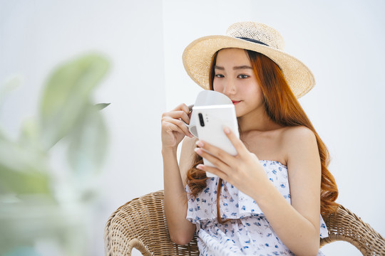 美丽的亚洲女旅行者戴着帽子，手持智能手机和杯子。